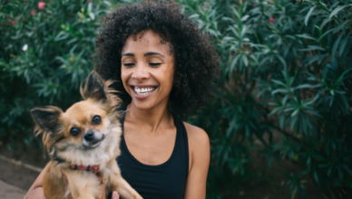 puppy owners - African American woman with black curly hair in a black tank top, smiling while holding her small tan puppy.