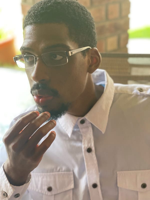 Author Jordan Spicer is looking off to the side with his right hand near his chin. He is wearing a with shirt with black buttons and wire rim glasses